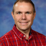 Smiling man with short hair wearing a red plaid shirt, set against a blue and purple background.