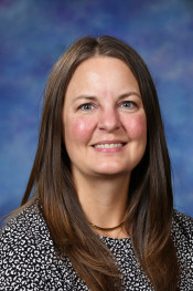 A smiling woman with long brown hair, wearing a black and white patterned shirt, against a blue background.
