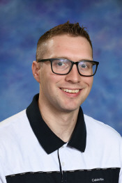 Smiling man with short hair and glasses, wearing a black and white collared shirt, against a blue background.