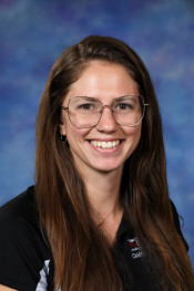 A smiling woman with long brown hair and glasses, wearing a black shirt, poses against a blue background.