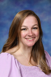 A woman with long brown hair smiles warmly, wearing a light purple blouse, against a soft blue background.