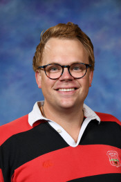 Smiling man with glasses wearing a black and red striped polo shirt, against a blue background.