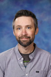 A man with a beard and short hair smiles in front of a blue background, wearing a gray shirt over a striped shirt.