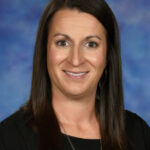 A woman with long brown hair smiles at the camera, wearing a black shirt against a blue background.