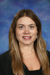 Headshot of a woman with long brown hair, wearing a black top, smiling against a blue background.