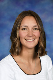 Smiling young woman with shoulder-length brown hair, wearing a white top, against a blue and purple background.