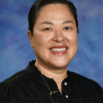 Smiling woman with dark hair, wearing a black blouse, against a blue and purple background.