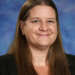 Portrait of a woman with long brown hair, smiling against a blue and purple background. She wears a black top.