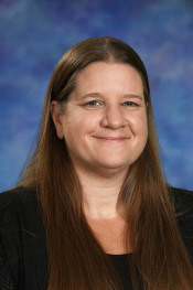 Portrait of a woman with long brown hair, smiling against a blue and purple background. She wears a black top.