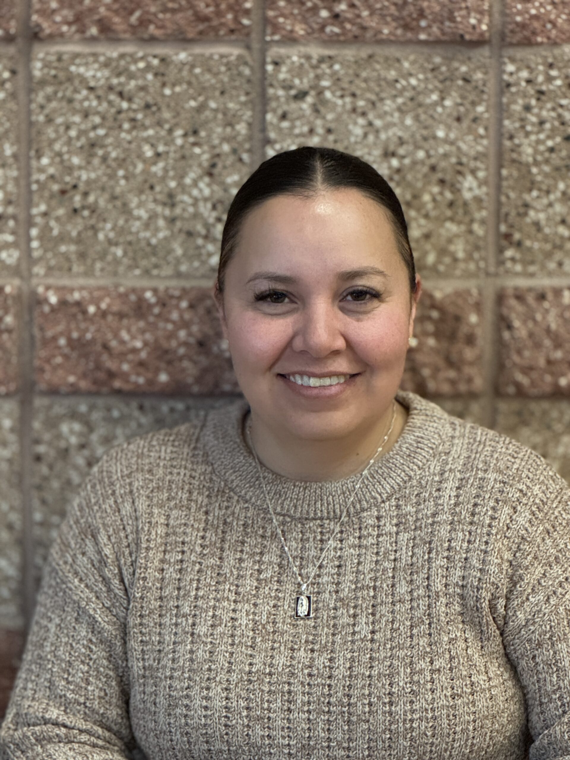 A smiling woman with dark hair in a bun wears a beige sweater, sitting against a textured stone wall.