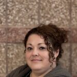 A woman with curly hair smiles, wearing a cozy sweater, against a textured brick wall background.
