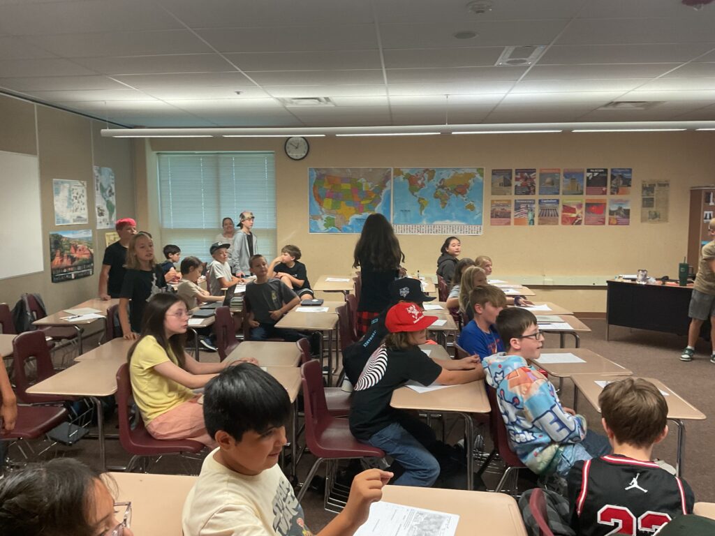 A classroom filled with students engaged in various activities, with maps and educational posters on the walls.