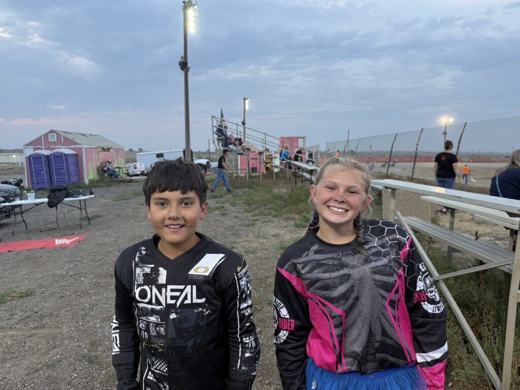 Two kids smiling in motocross gear at an outdoor event, with bleachers and spectators in the background.