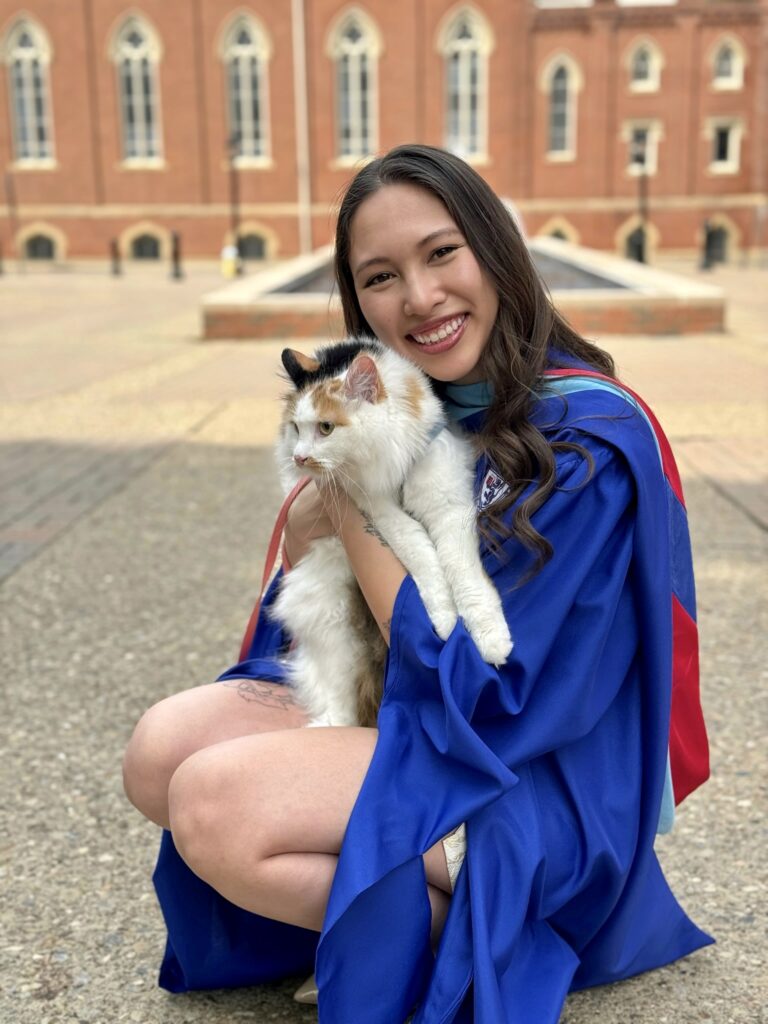 A graduate in a blue gown smiles while holding a fluffy cat, with a brick building and fountain in the background.