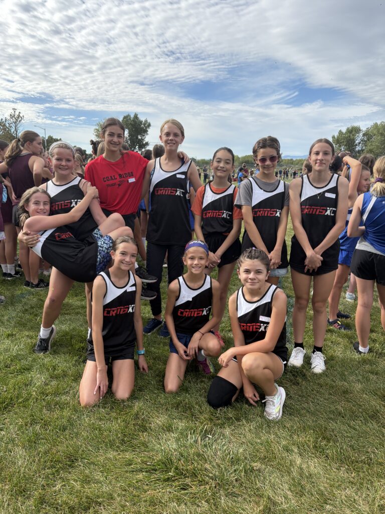 A group of young female athletes in running uniforms pose together on grass, smiling and ready for a race.