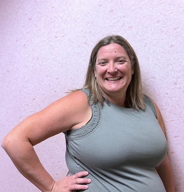 Smiling woman with long hair poses with one hand on her hip against a light pink textured wall.