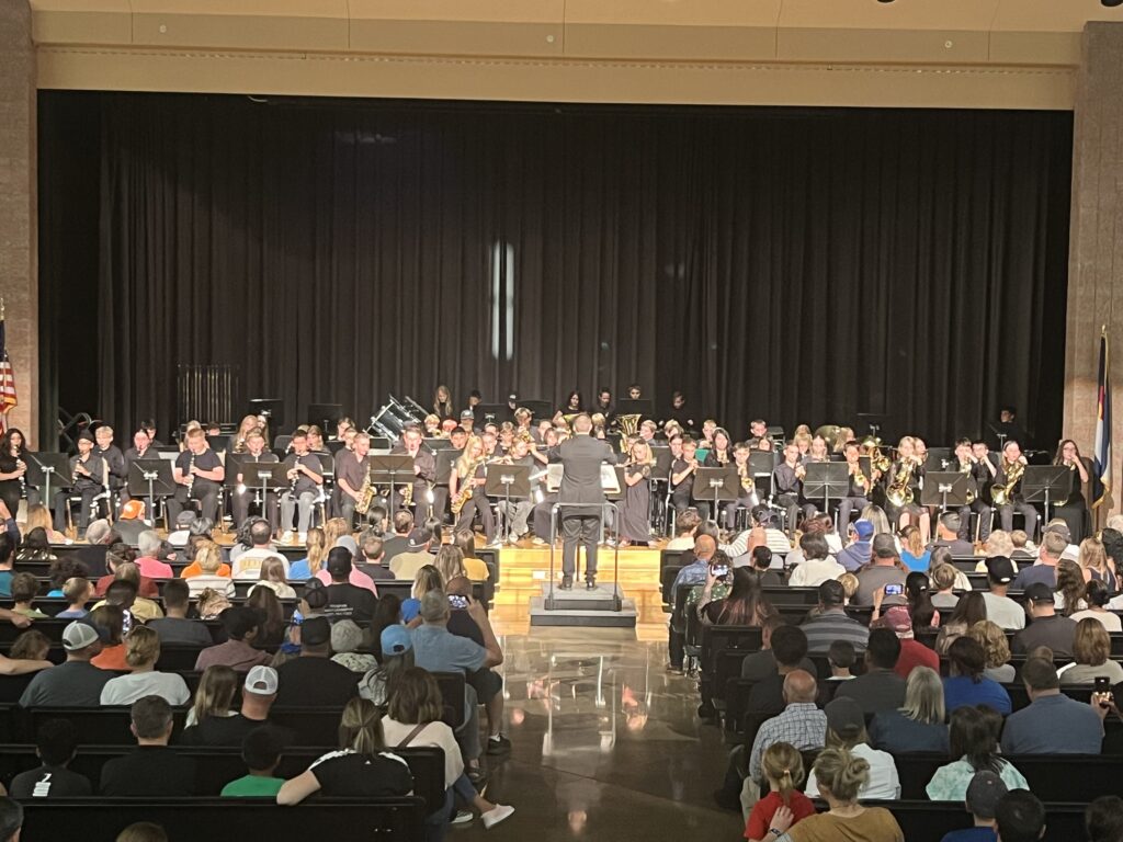 A large band performs on stage in front of an audience, with musicians playing various instruments.