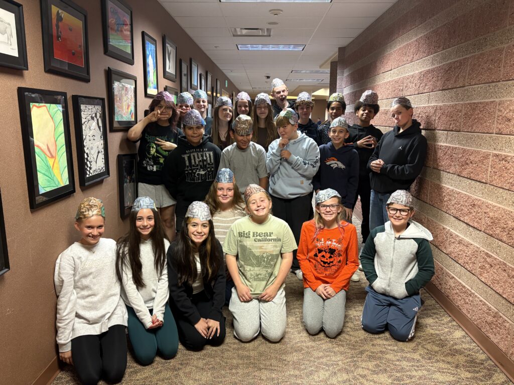 A group of children and teens wearing foil hats pose together in a hallway decorated with artwork.
