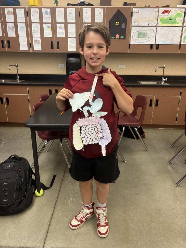 A boy holds a paper model of human organs, smiling in a classroom with drawings on the walls behind him.