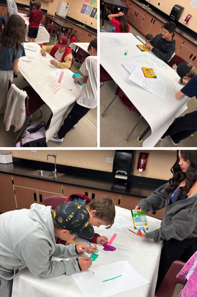 Students are engaged in art projects on a large table covered with paper, using various art supplies in a classroom.