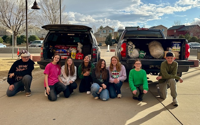 A group of eight people pose in front of two SUVs filled with donations, smiling on a sunny day.