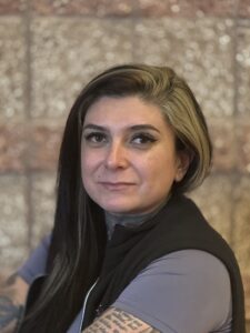 A person with long black and blonde hair, wearing a dark vest, poses with arms crossed against a textured background.