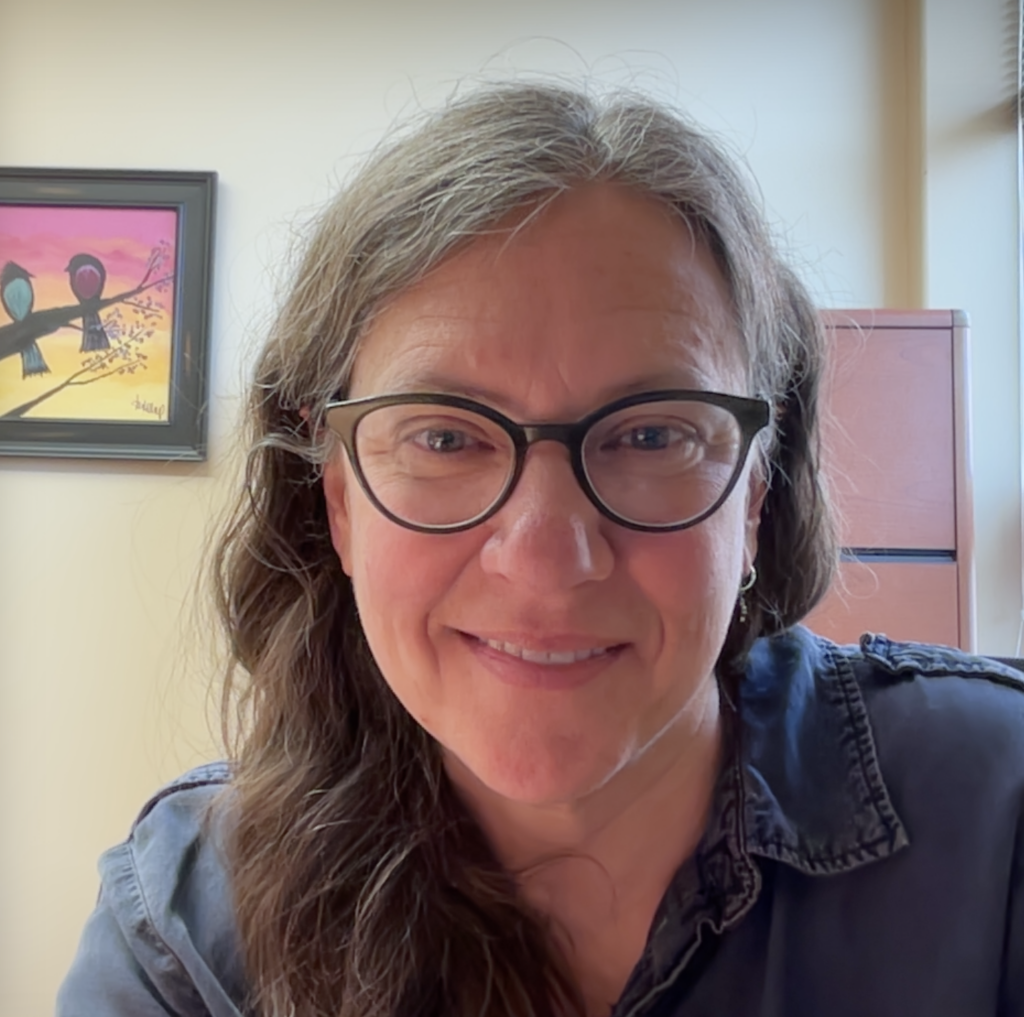 A smiling woman with long hair and glasses sits in front of a colorful bird painting in an office setting.