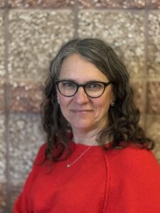 A woman with curly hair and glasses smiles, wearing a red sweater against a textured wall background.