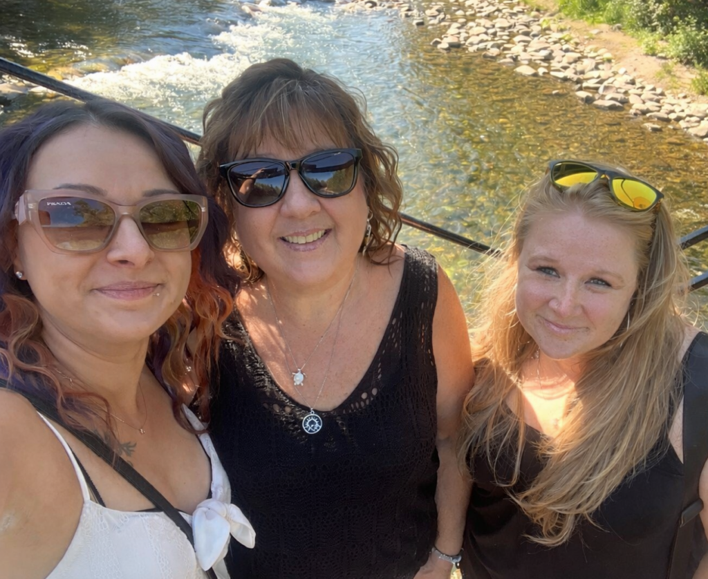 Three women smile for a selfie by a river with rocks, greenery, and sunlight in the background.