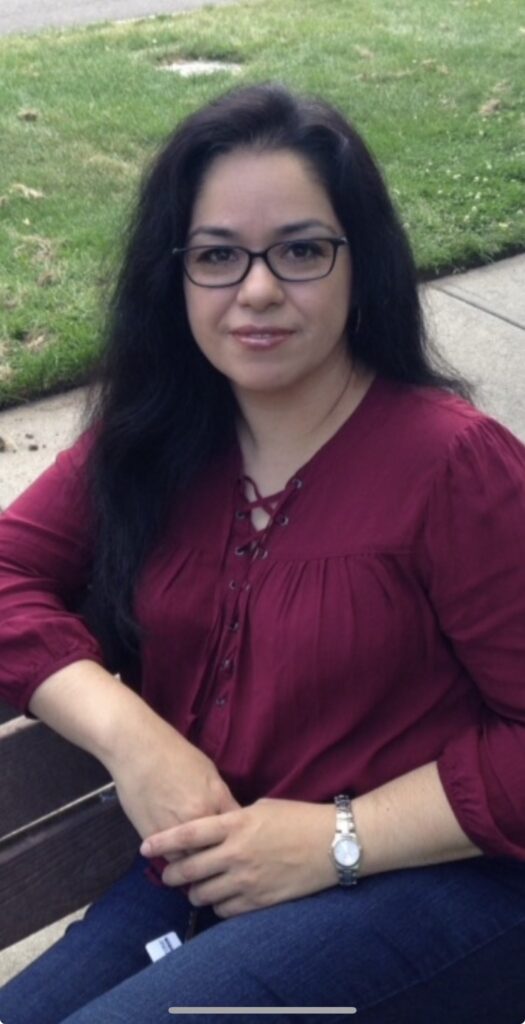 A woman with long dark hair and glasses sits on a bench, wearing a burgundy blouse and jeans, smiling softly.