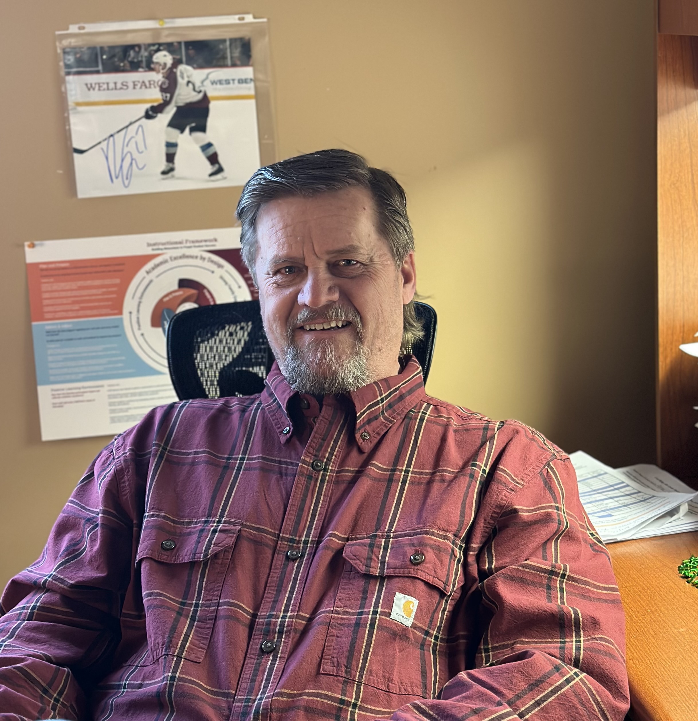 A smiling man with a beard sits in an office chair, wearing a plaid shirt, with documents and a signed sports photo in the background.