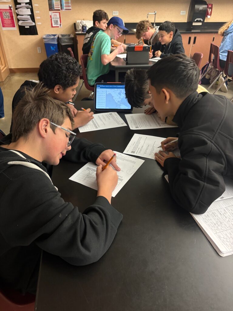 Students collaborate at a table, working on papers while using a tablet for research in a classroom setting.