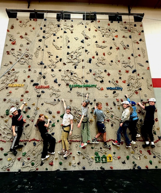 Eight children in helmets climb a rock wall with values like "Respect" and "Honesty" displayed above them.