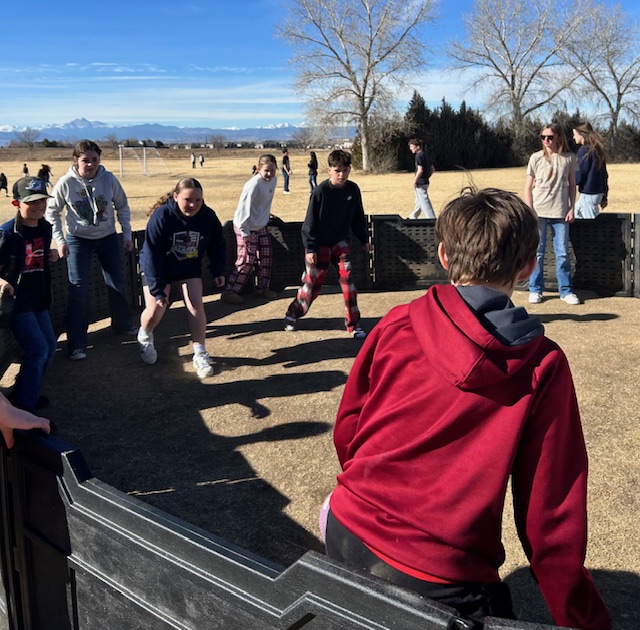 A group of children playing a game outside on a sunny day, with mountains in the background and a grassy field.