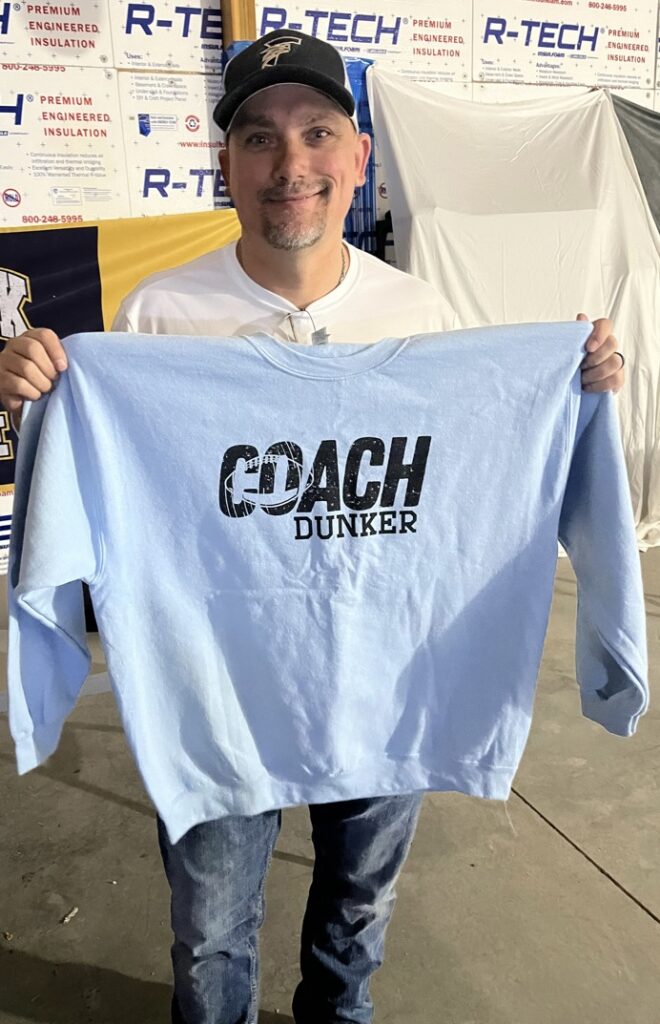 A man holds up a light blue sweatshirt with "COACH DUNKER" printed on it, smiling in a casual setting.