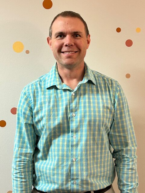 Man smiling in a light blue and green checked shirt, standing against a wall with colorful circular decorations.