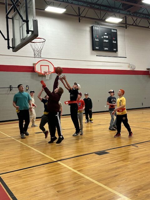 A group of children playing basketball in a gym, with one jumping to make a shot while others watch.