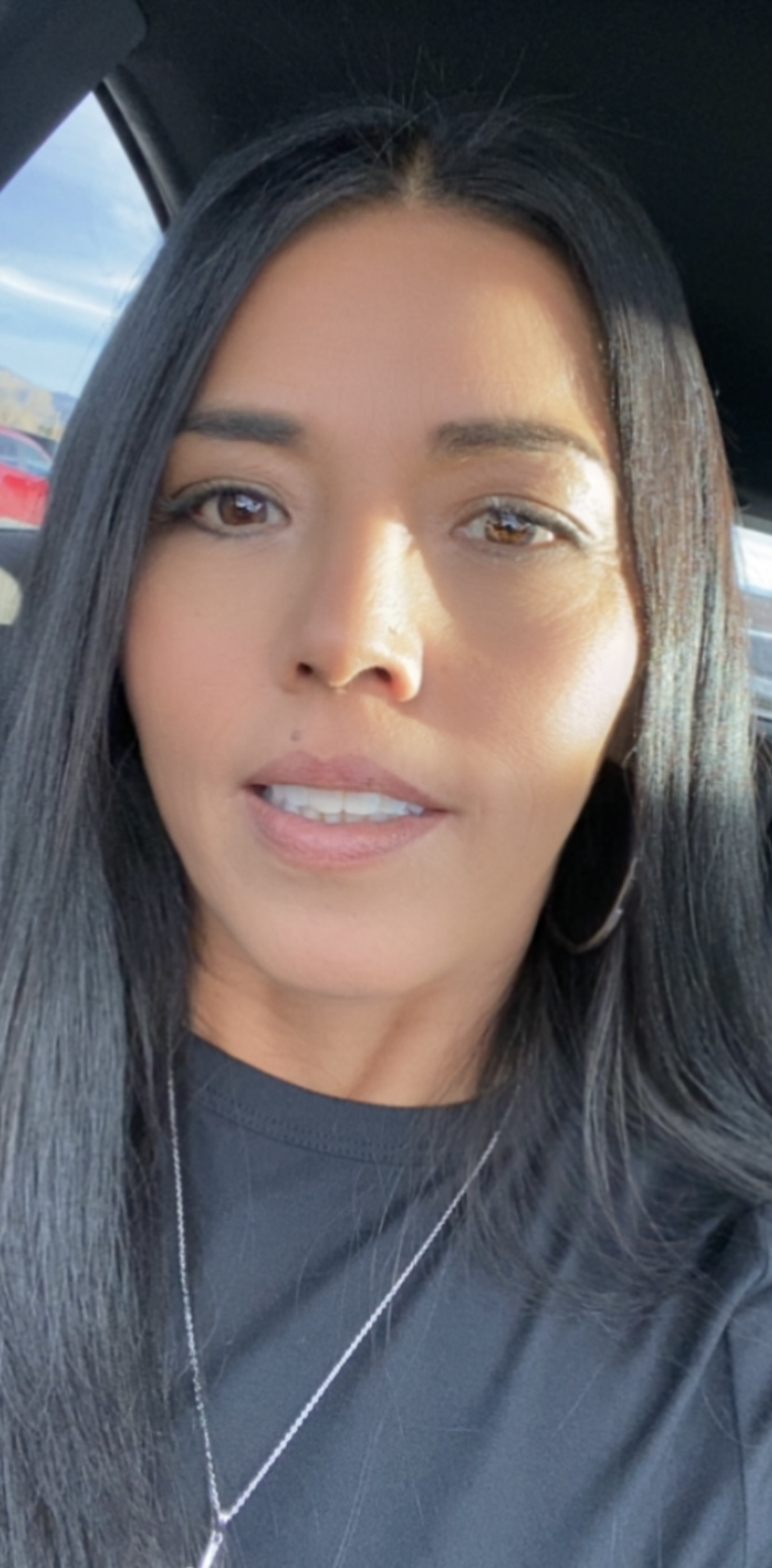 A woman with long black hair and hoop earrings smiles, sitting in a car with a bright background.