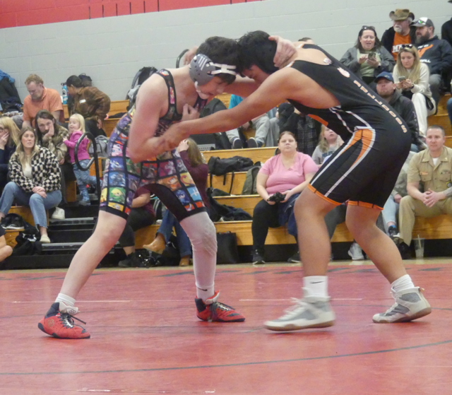 Two high school wrestlers are competing on a red mat, while spectators watch from the bleachers in the background.
