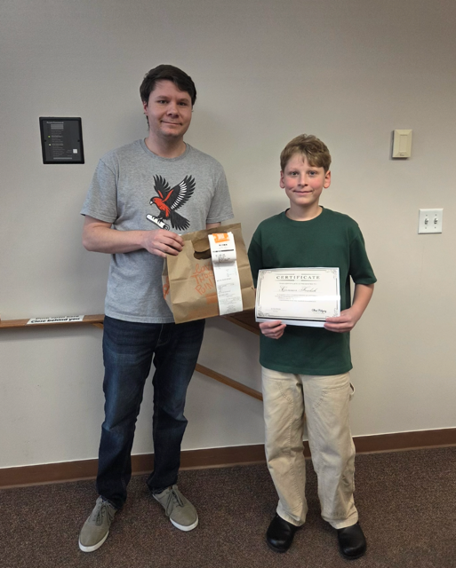 A man and a boy stand together, the man holding a bag and the boy holding a certificate, in a room with beige walls.