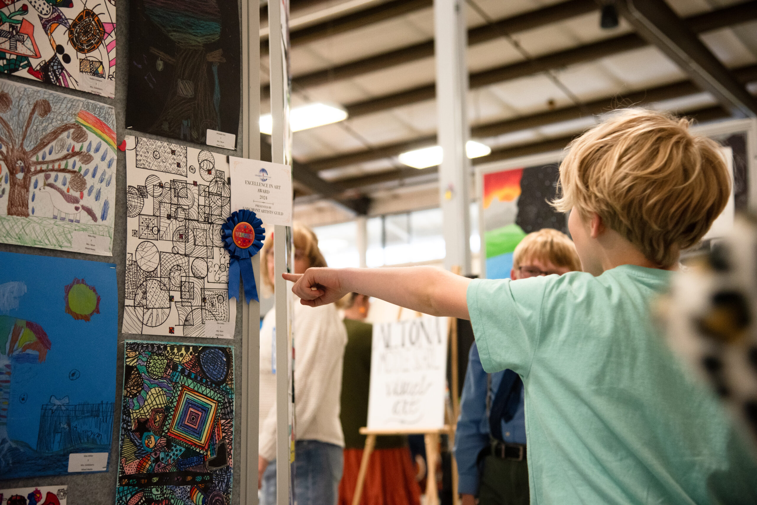 A child points at an award-winning artwork displayed at an art show, surrounded by other attendees.