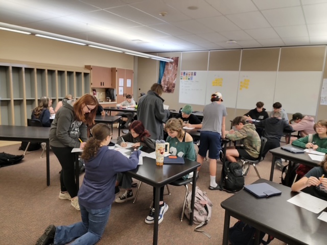 A classroom with students engaged in group work at tables, some writing, and others collaborating on projects.