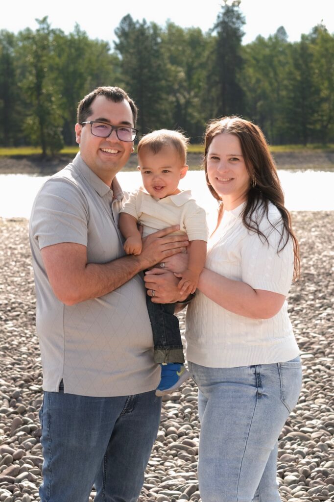 A smiling family of three stands on a rocky riverbank, with trees and water in the background. The child is held by the father.