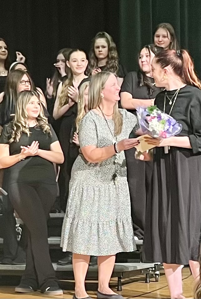 A woman receives a bouquet and award from another woman, while a group of girls in black dresses applauds in the background.