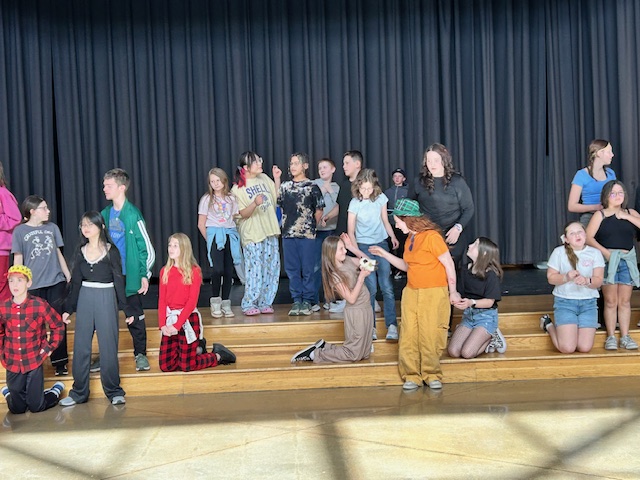 A group of students poses on stage, dressed in various outfits, with a black curtain in the background.