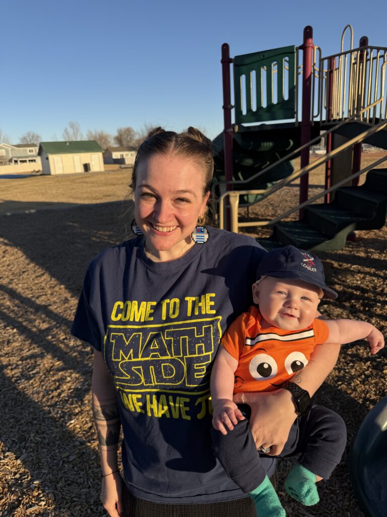 A woman smiles while holding a baby in a playground, both wearing colorful outfits and enjoying the sunny day.