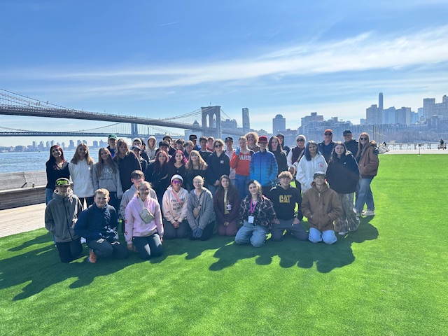 A large group of students poses on green grass near the Brooklyn Bridge and skyline on a sunny day.