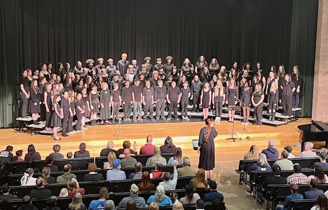 A large choir performs on stage in black attire, while an audience watches from below.