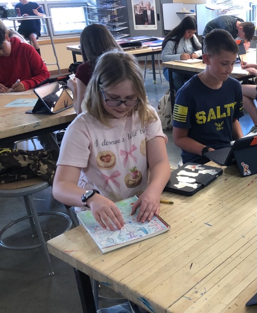 A student with glasses sits at a table, coloring in a sketchbook while others work on tablets in a classroom.