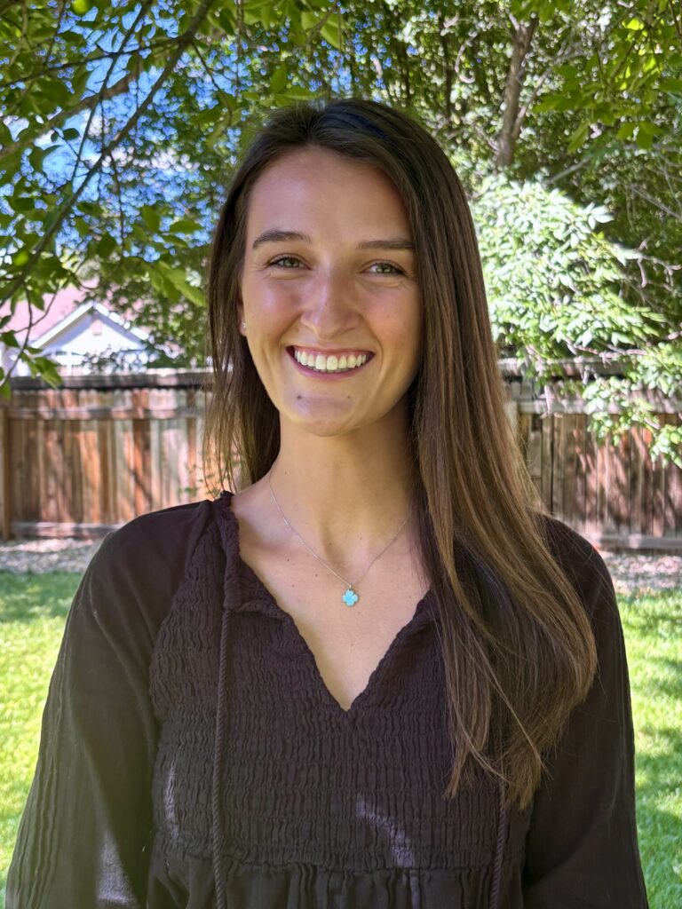 A young woman with long brown hair smiles outdoors, wearing a black top and a turquoise necklace, surrounded by greenery.